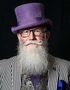 Portrait of a bearded man with a curled moustache wearing a purple top hat and a pair of glasses and a black and pale grey striped jacket. His name is David Dade honorary president of the British Beard Club.